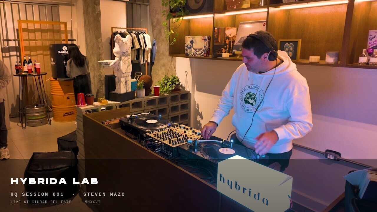 DJ Steven Mazo at the decks inside HYBRIDA LAB HQ, wearing the Atelier Hybrida hoodie. The HYBRIDA wordmark lightbox sits in the foreground.